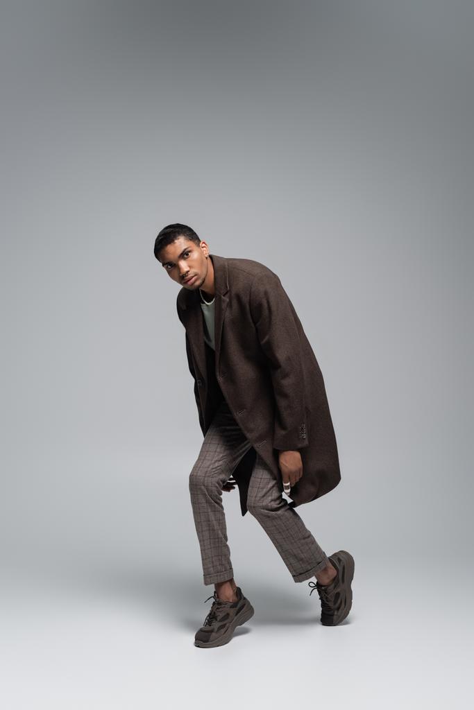 in voller Länge junger afrikanisch-amerikanischer Mann in herbstlichem Wollmantel, der in die Kamera schaut und auf grau posiert  - Foto, Bild
