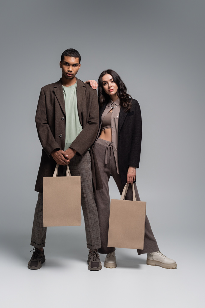 full length of stylish interracial couple in trendy outfits holding shopping bags on grey - Photo, Image
