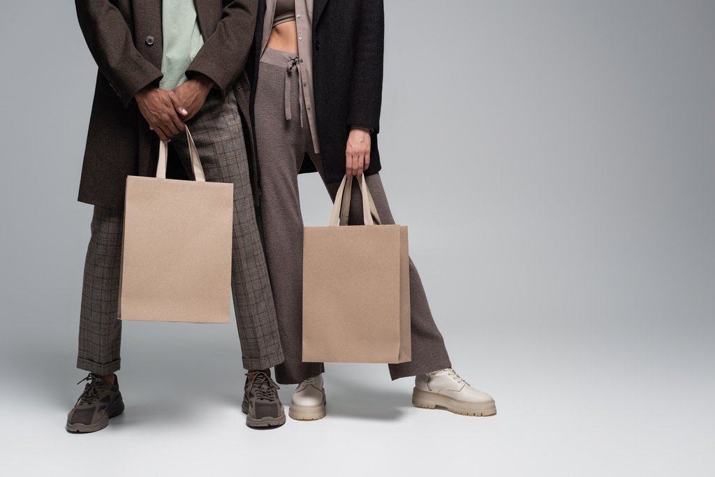 vue recadrée du couple interracial élégant dans des tenues automnales tenant des sacs à provisions sur gris - Photo, image