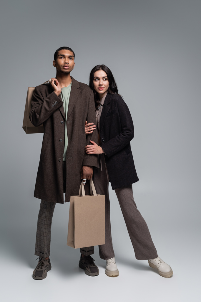 longitud completa de elegante pareja interracial en trajes otoñales de moda sosteniendo bolsas de compras en gris - Foto, Imagen