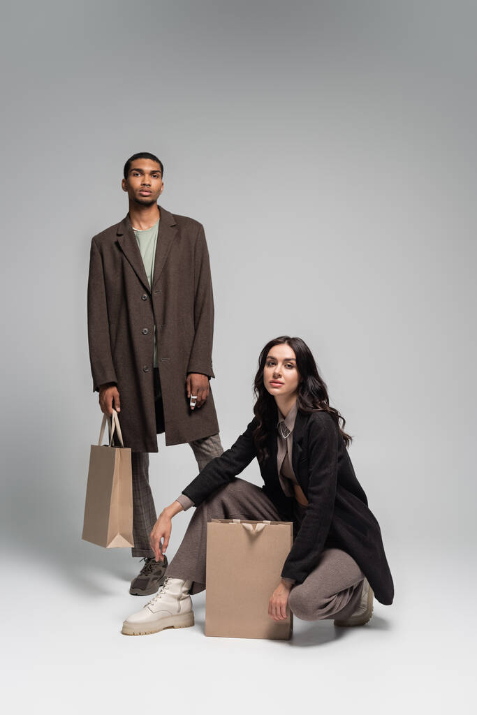 full length of stylish interracial couple in autumnal outfits posing near paper bags on grey - Photo, Image