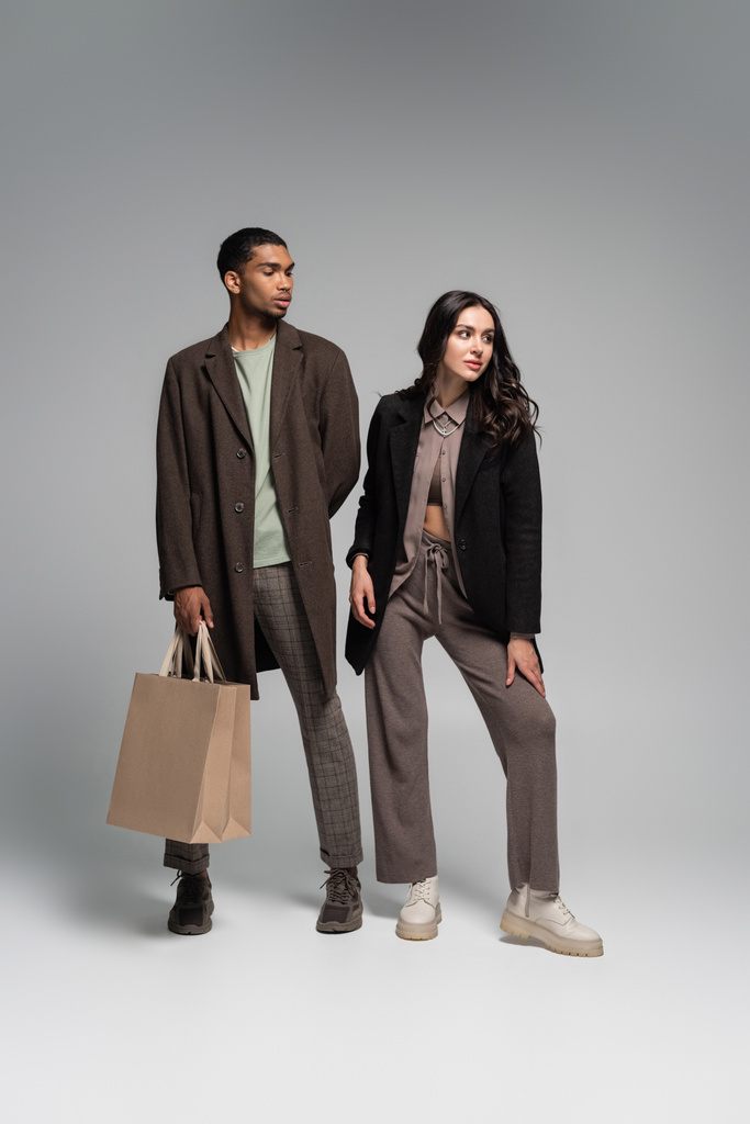 full length of stylish african american in autumnal coat posing with paper bags near woman on grey - Photo, Image