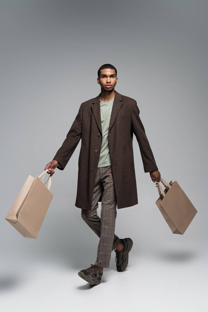 longueur totale de l'homme afro-américain élégant en manteau de laine et baskets posant avec des sacs à provisions sur gris - Photo, image