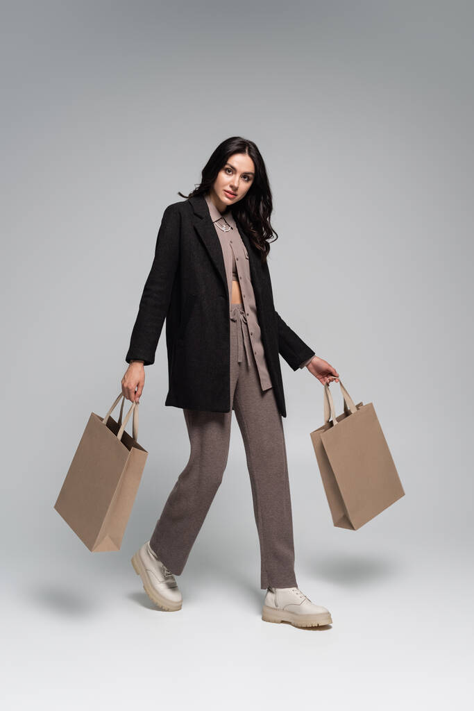 pełna długość młodej kobiety w stylowym jesiennym stroju spaceru z torbami na zakupy na szarości - Zdjęcie, obraz