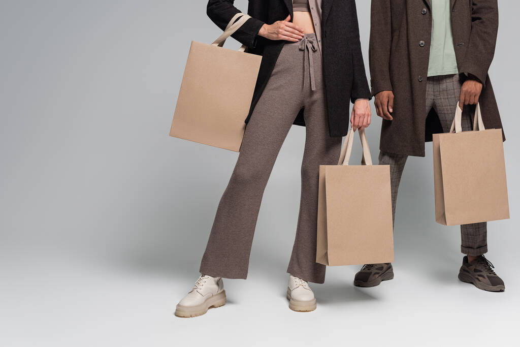 partial view of stylish interracial couple in autumnal outfits holding shopping bags on grey - Photo, Image