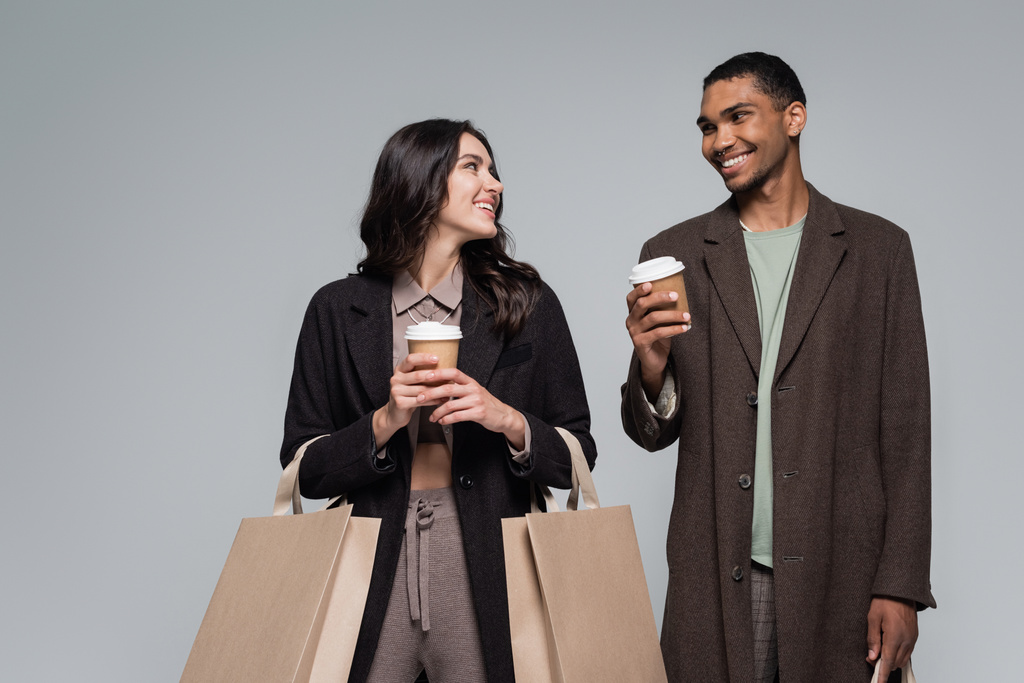 felice coppia interrazziale in abiti eleganti che tengono borse della spesa e bicchieri di carta isolati su grigio - Foto, immagini