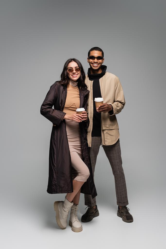 pleine longueur de couple interracial heureux dans des tenues élégantes et des lunettes de soleil tenant des tasses en papier sur gris - Photo, image