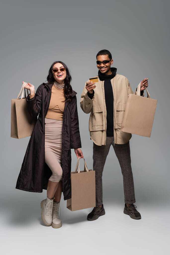 in voller Länge glückliche interrassische Models in stylischer Sonnenbrille mit Einkaufstaschen und Kreditkarte auf grau - Foto, Bild