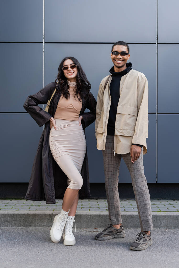 полная длина стильной межрасовой пары в осенних нарядах и солнцезащитных очках, стоящих рядом со зданием  - Фото, изображение
