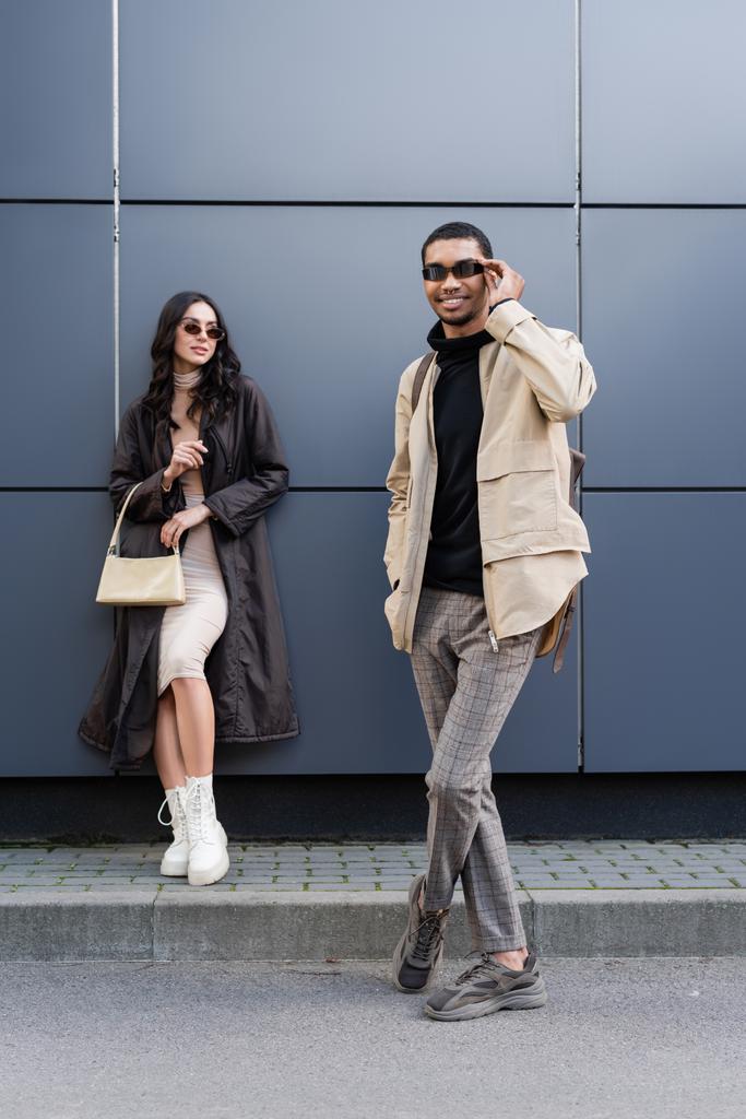 full length of happy african american man adjusting sunglasses near stylish woman standing with handbag  - Photo, Image