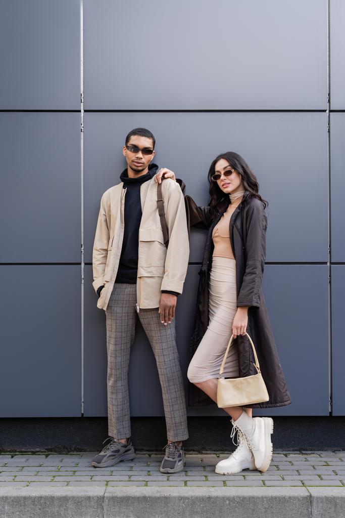 longitud completa de elegante pareja interracial en trajes otoñales y gafas de sol de moda de pie cerca del edificio  - Foto, Imagen