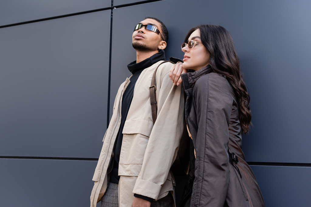 pareja interracial con estilo en trajes otoñales y gafas de sol de moda de pie cerca del edificio  - Foto, Imagen