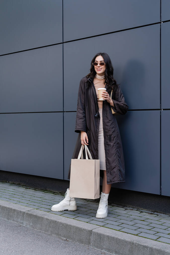 pełna długość szczęśliwy młoda kobieta w stylowy strój i okulary przeciwsłoneczne trzymając papierowy kubek i torbę na zakupy w pobliżu budynku  - Zdjęcie, obraz