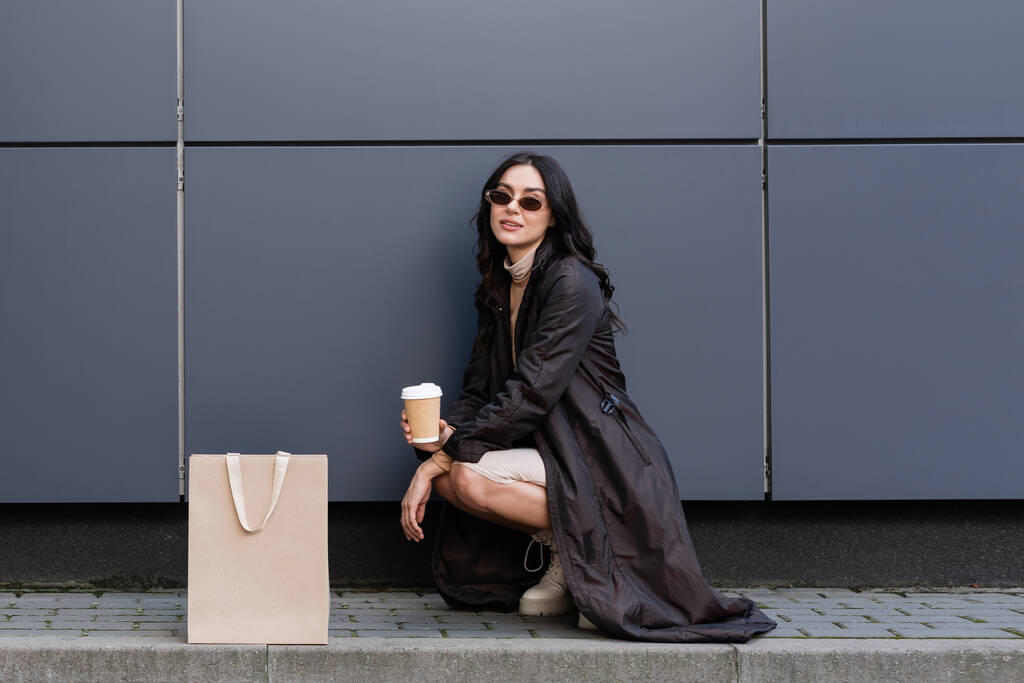 young stylish woman with paper cup sitting near paper bag and building of shopping mall  - Photo, Image