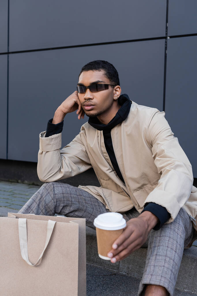 africký Američan muž v slunečních brýlích a podzimní oblečení sedí s kávou jít do blízkosti nákupní tašky  - Fotografie, Obrázek