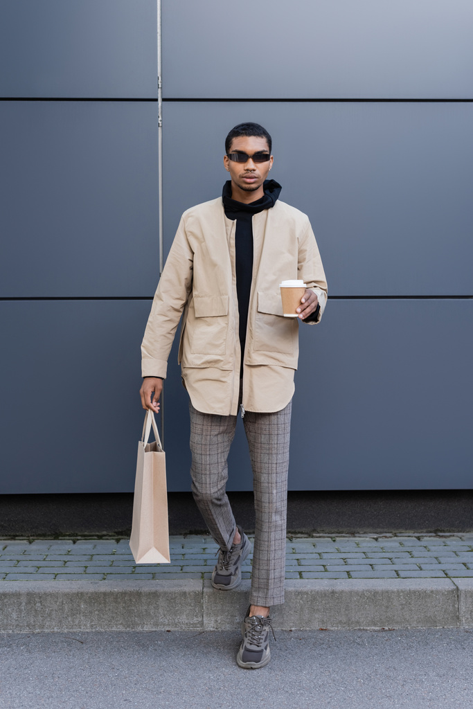 longitud completa de hombre afroamericano con estilo en gafas de sol y traje otoñal sosteniendo taza de papel y bolsa de compras  - Foto, Imagen