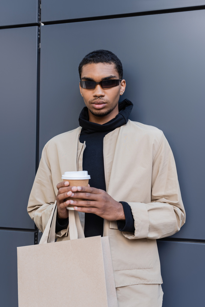 homem americano africano elegante em óculos de sol e outfit outnal segurando copo de papel e saco de compras  - Foto, Imagem