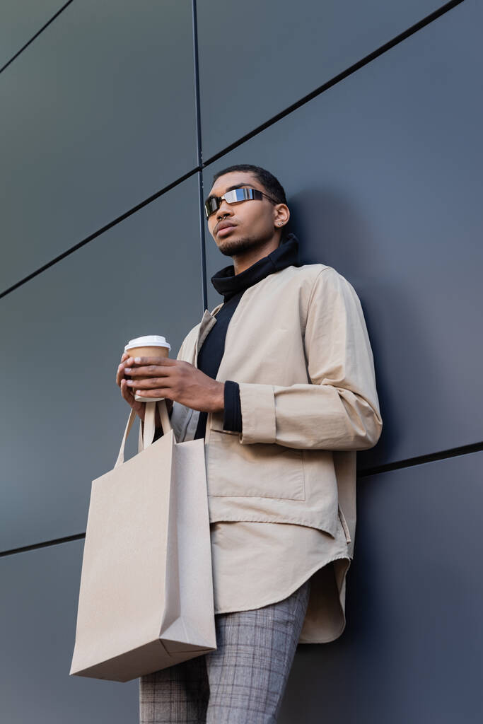 niski kąt widzenia stylowy Afroamerykanin człowiek w okularach przeciwsłonecznych i strój jesienny trzymając papierowy kubek i torbę na zakupy  - Zdjęcie, obraz