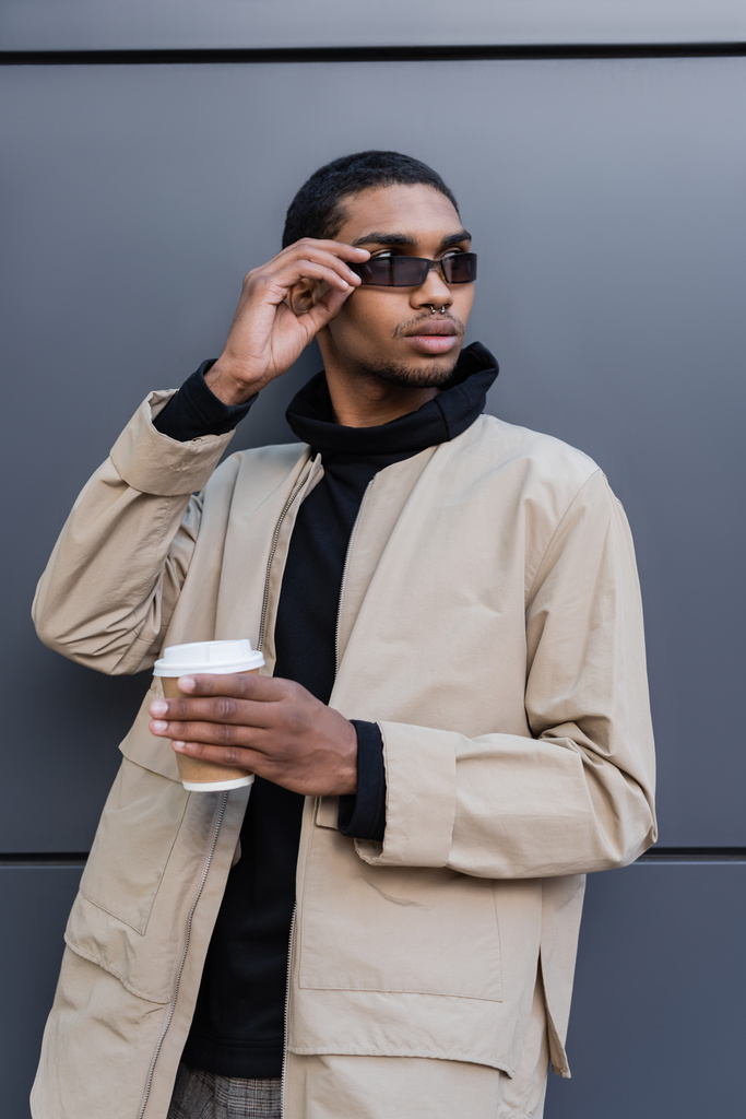 elegante uomo africano americano in abito autunnale che tiene tazza di carta e occhiali da sole di regolazione - Foto, immagini