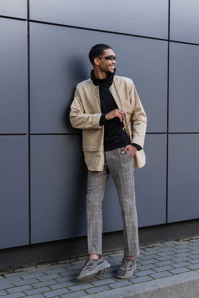 felice uomo africano americano in abito autunnale e occhiali da sole alla moda in piedi con mano in tasca vicino edificio - Foto, immagini