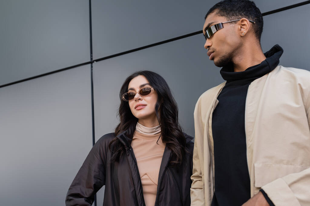 низкий угол обзора стильной многонациональной пары в модных осенних нарядах и солнцезащитных очках, стоящих рядом со зданием  - Фото, изображение