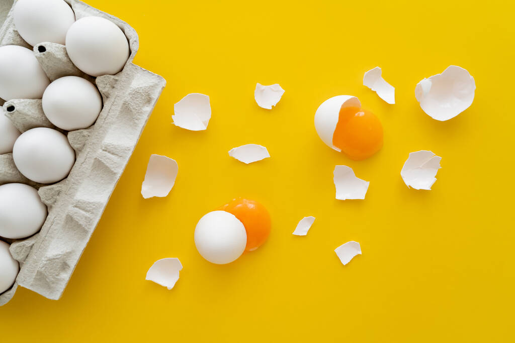 Bovenaanzicht van rauwe dooiers en gebarsten schelpen in de buurt van eieren in dienblad op gele achtergrond  - Foto, afbeelding