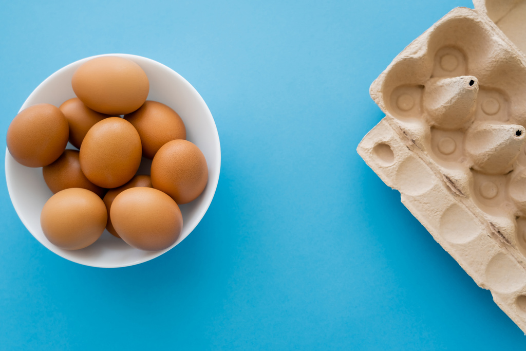 Horní pohled na karton kontejner a kuřecí vejce v misce na modrém pozadí - Fotografie, Obrázek
