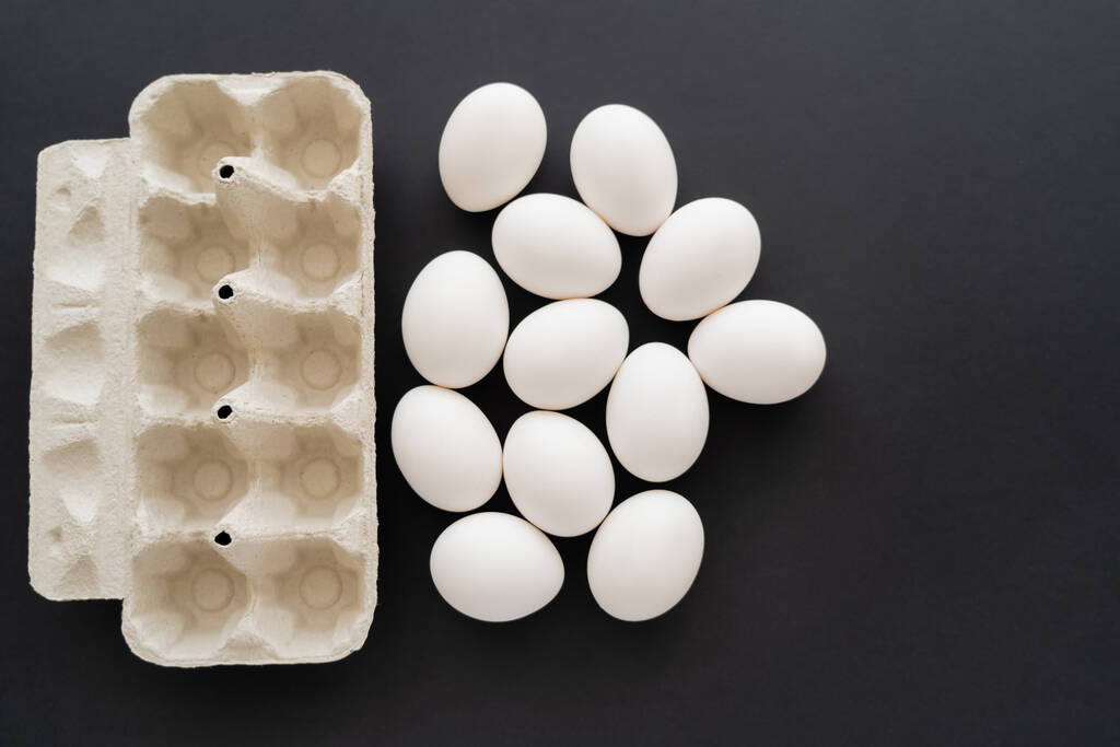 Vista dall'alto di uova di pollo bianche vicino al contenitore di cartone su sfondo nero  - Foto, immagini