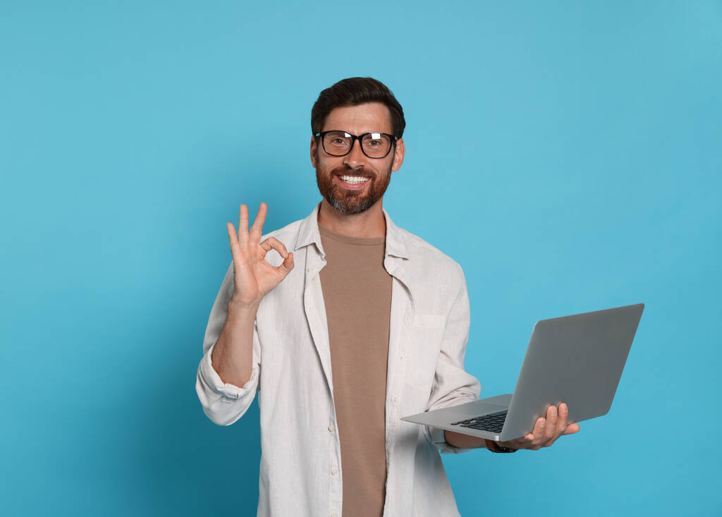 Schöner Mann mit Laptop zeigt OK-Geste auf hellblauem Hintergrund - Foto, Bild