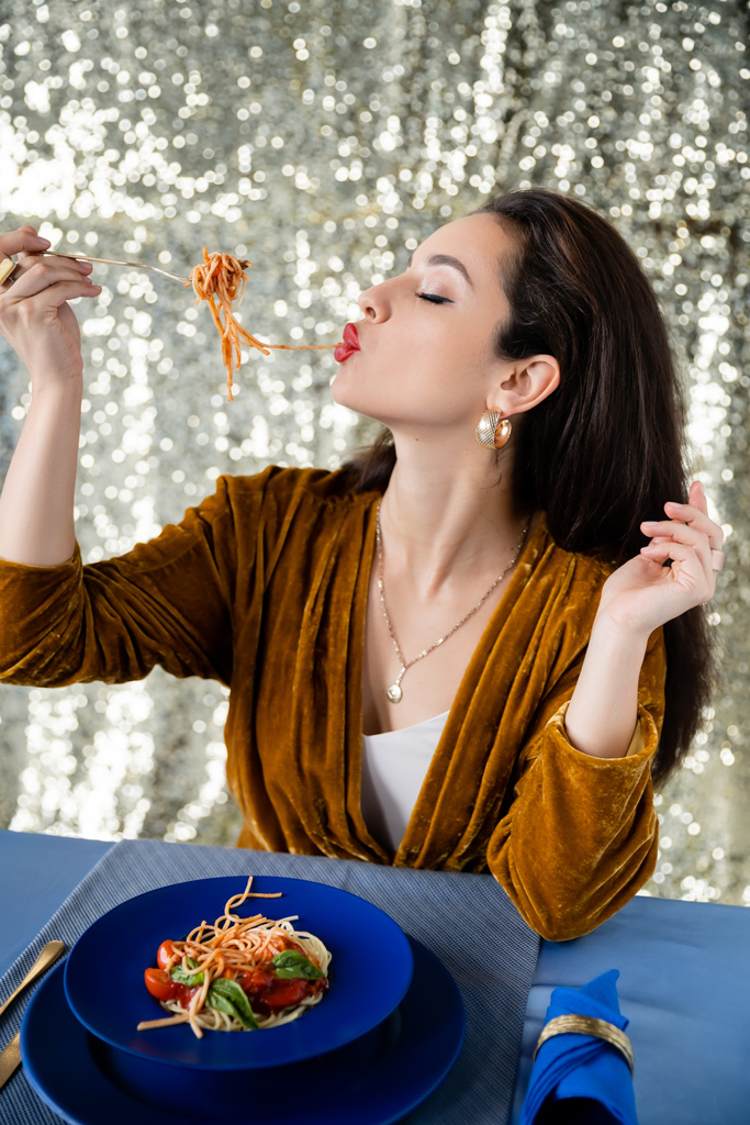 brunette woman eating tasty spaghetti near blue plates on shiny silver background - Photo, Image