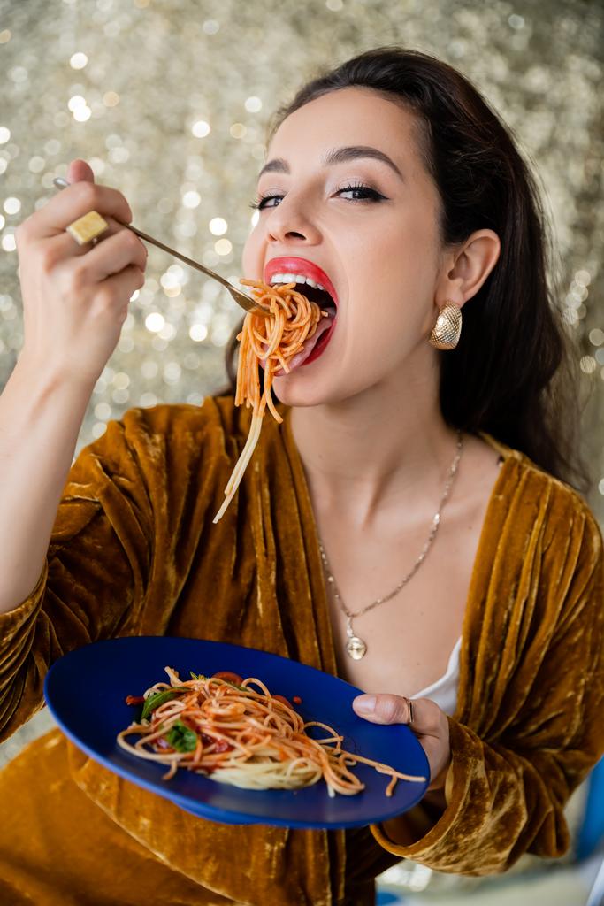 женщина в велюровом платье ест вкусные спагетти и смотрит в камеру на блестящем серебряном фоне - Фото, изображение