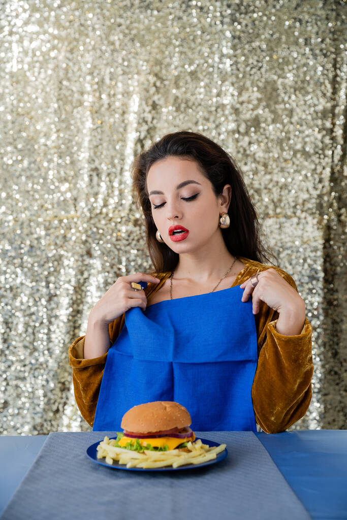 svůdná žena drží modrý ubrousek u talíře s hranolky a burger na lesklém stříbrném pozadí - Fotografie, Obrázek