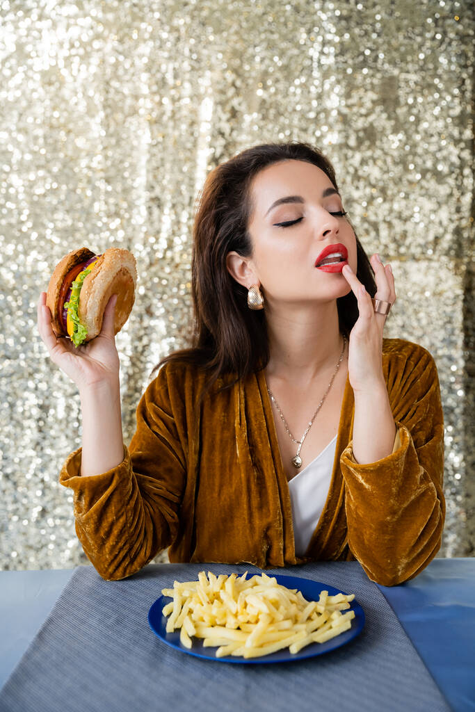 Gözleri kapalı, dudağına dokunan ve patates kızartmasının yanında hamburger tutan baştan çıkarıcı bir kadın. - Fotoğraf, Görsel