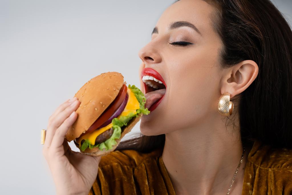 elegáns fiatal nő smink eszik finom burger elszigetelt szürke - Fotó, kép