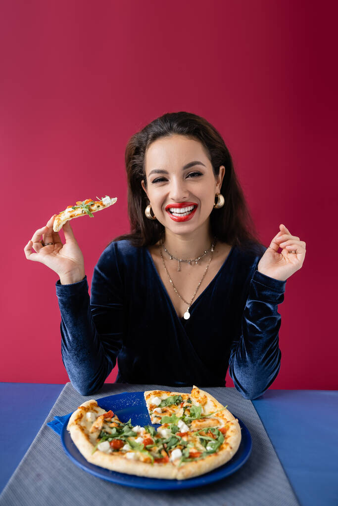 χαρούμενη γυναίκα σε βελούδινο φόρεμα κρατώντας πίτσα και κοιτάζοντας κάμερα κοντά στο μπλε τραπέζι που απομονώνονται σε κόκκινο - Φωτογραφία, εικόνα
