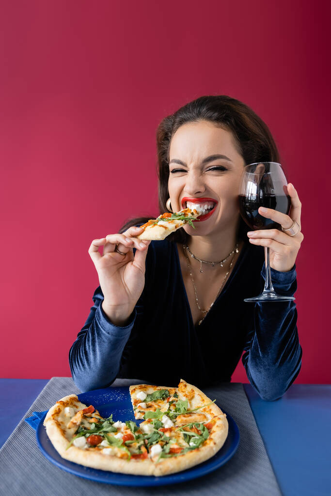 fröhliche Frau mit einem Glas Wein, die leckere Pizza in der Nähe des blauen Tisches isst, isoliert auf rot - Foto, Bild