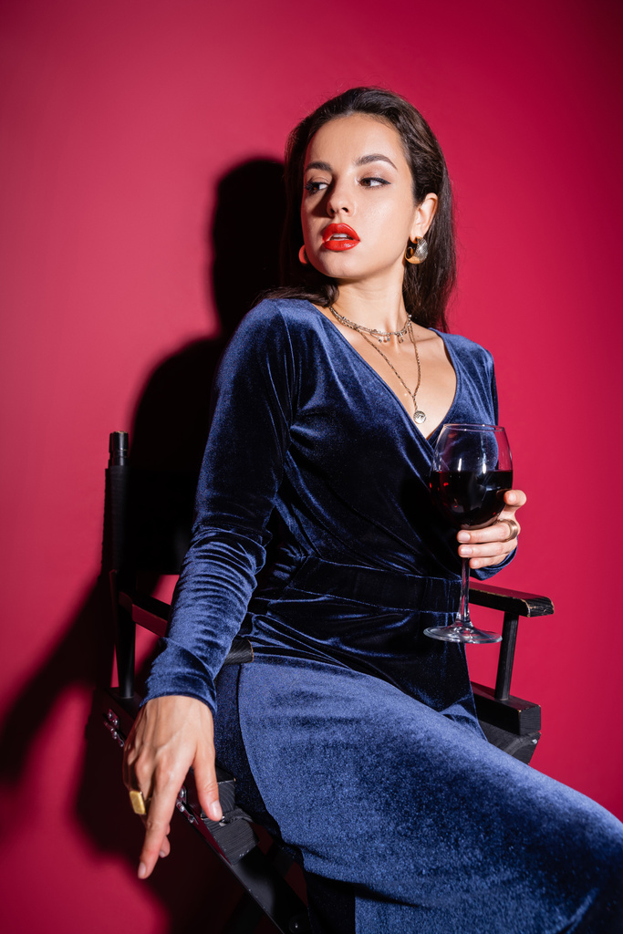 σαγηνευτική γυναίκα σε κομψό φόρεμα κάθεται με ένα ποτήρι κρασί και κοιτάζοντας μακριά στο κόκκινο φόντο - Φωτογραφία, εικόνα