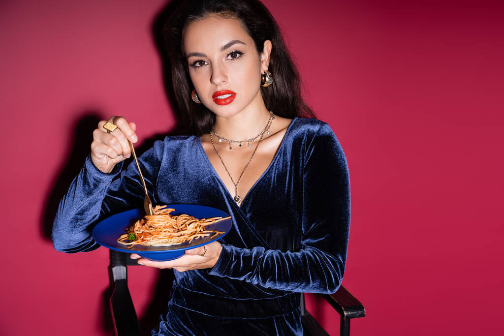 sensual woman in elegant dress holding blue plate with pasta and looking at camera on red background - Photo, Image