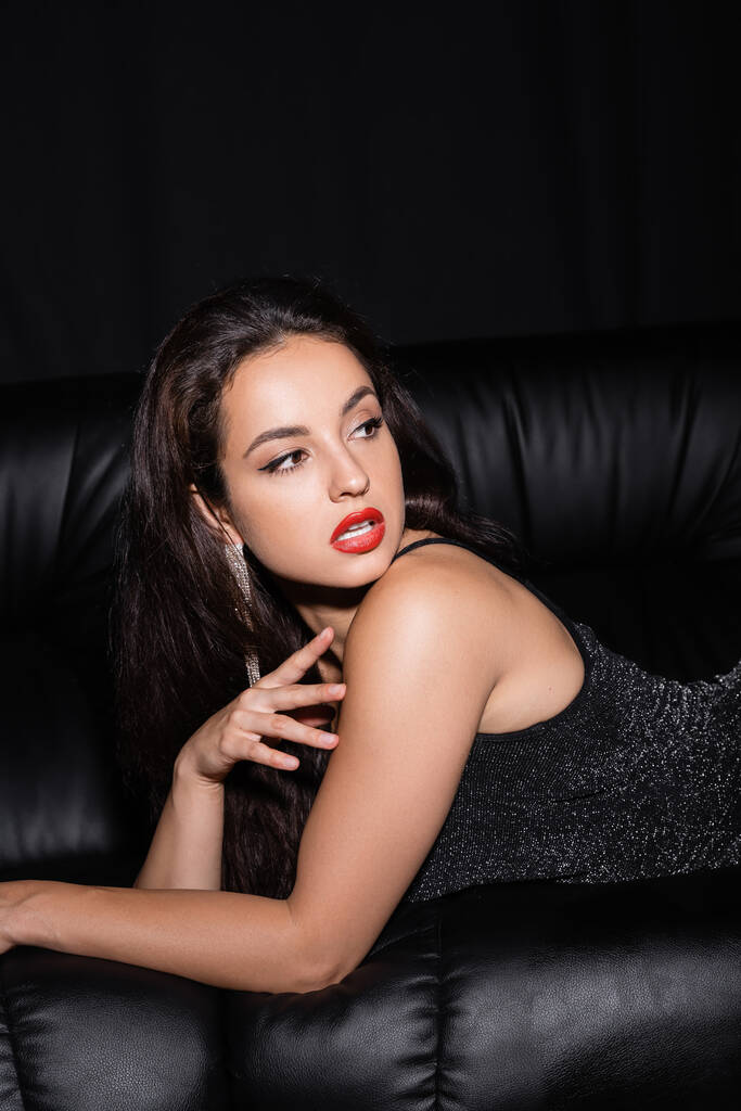 donna glamour con labbra rosse guardando altrove mentre giaceva sul divano di pelle isolato su nero - Foto, immagini