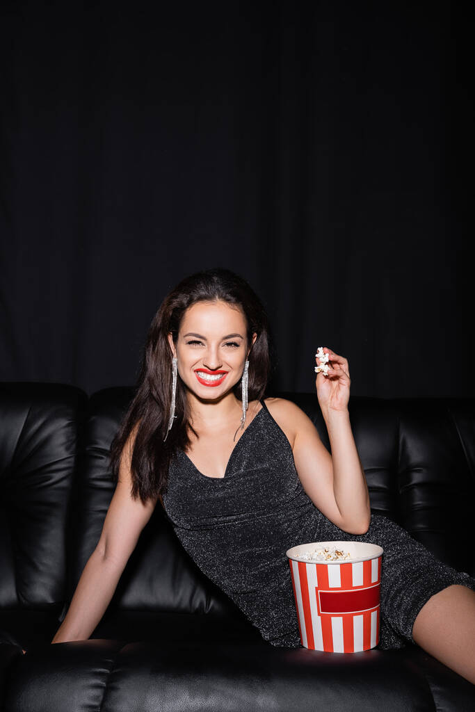 donna allegra guardando macchina fotografica vicino secchio di carta di popcorn isolato su nero - Foto, immagini
