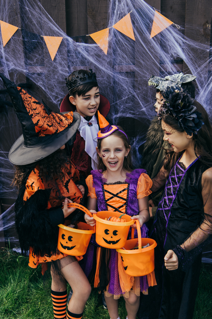 Cheerful girl in costume holding bucket with candies near interracial friends and spider web on fence during halloween party - Photo, Image