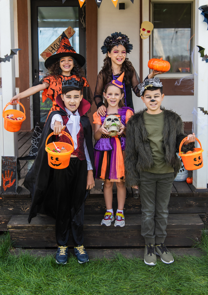 cheerful multiethnic friends in halloween costumes holding buckets with candies near decorated cottage - Photo, Image