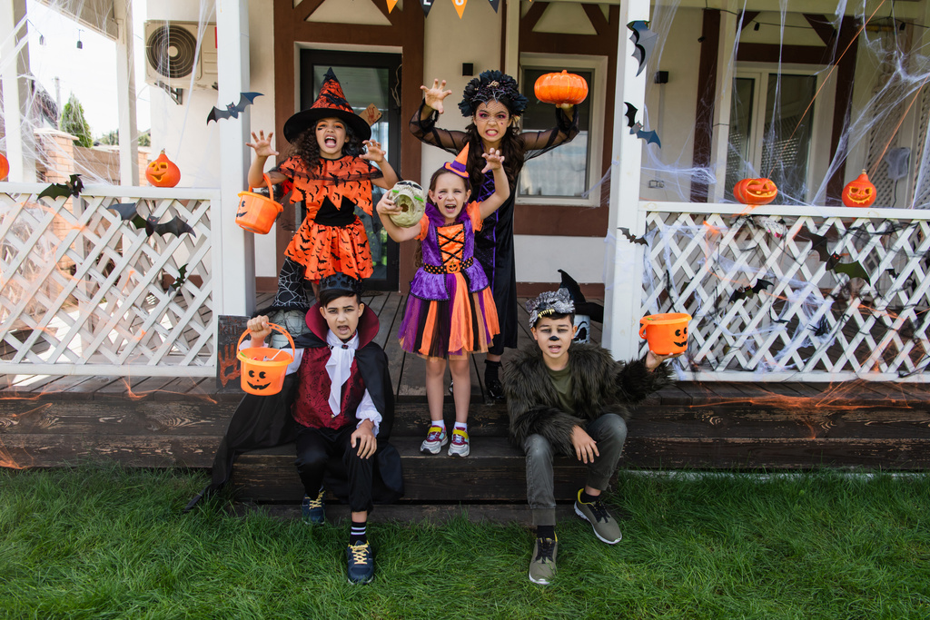 niños multiétnicos en disfraces de Halloween haciendo muecas y mostrando gestos de miedo cerca de casa de campo decorada - Foto, Imagen