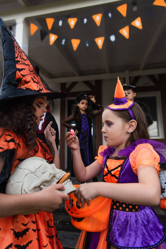 ragazze in costumi di Halloween con teschio e secchio con caramelle vicino sfocati amici multietnici - Foto, immagini