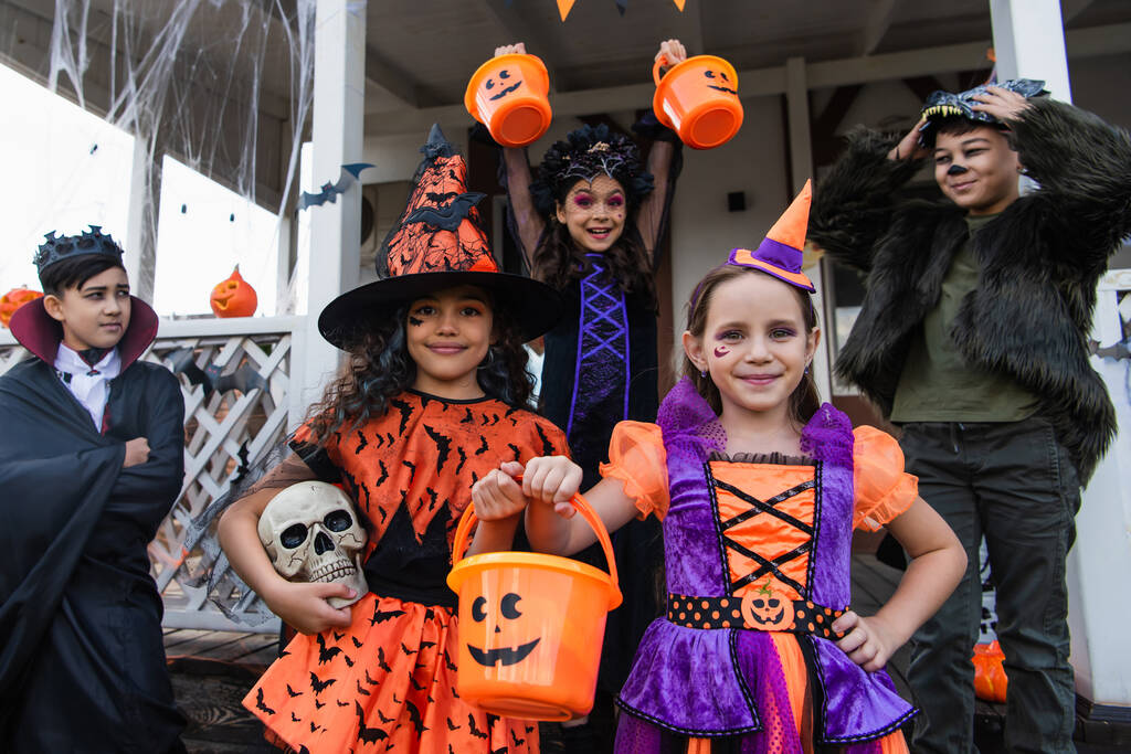 alegre interracial niños en halloween trajes de pie con truco o tratar cubos cerca de casa - Foto, imagen