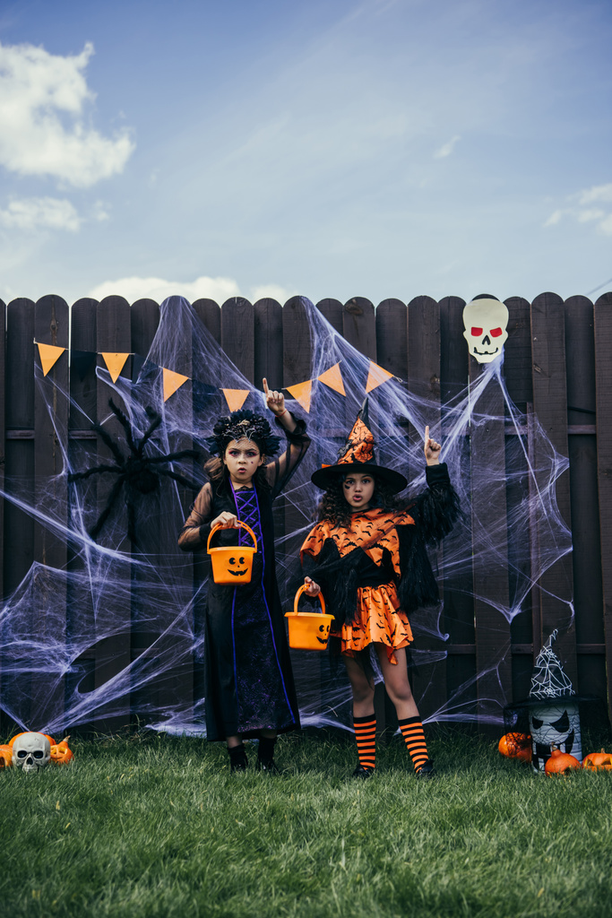 Meisjes in halloween kostuums houden emmers en wijzen met vingers in de buurt decor op hek buiten - Foto, afbeelding