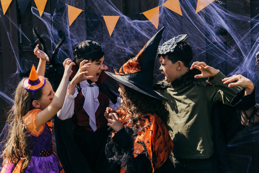 Międzyrasowe dzieci w kostiumach gryzące się na siebie w pobliżu wystroju halloween na podwórku - Zdjęcie, obraz