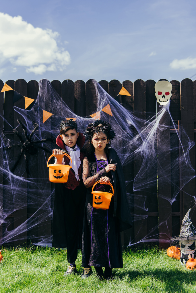 Multiethnische Kinder in Halloween-Kostümen halten Eimer in der Nähe von Dekoration auf Zaun und Gras im Freien - Foto, Bild