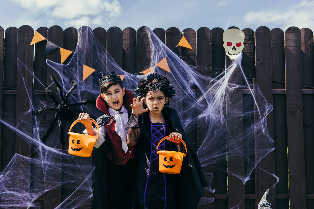 Interracial crianças em trajes segurando baldes halloween perto de teia de aranha em cerca ao ar livre - Foto, Imagem