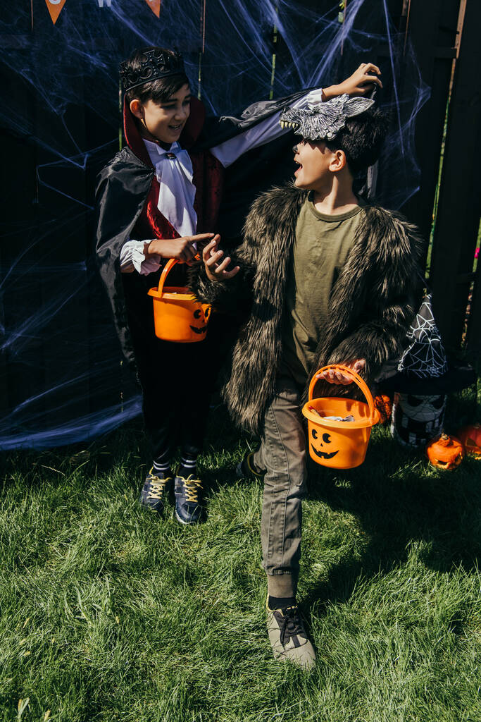 Cheerful asian boys in halloween costumes holding buckets with candies on grass in backyard - Photo, Image